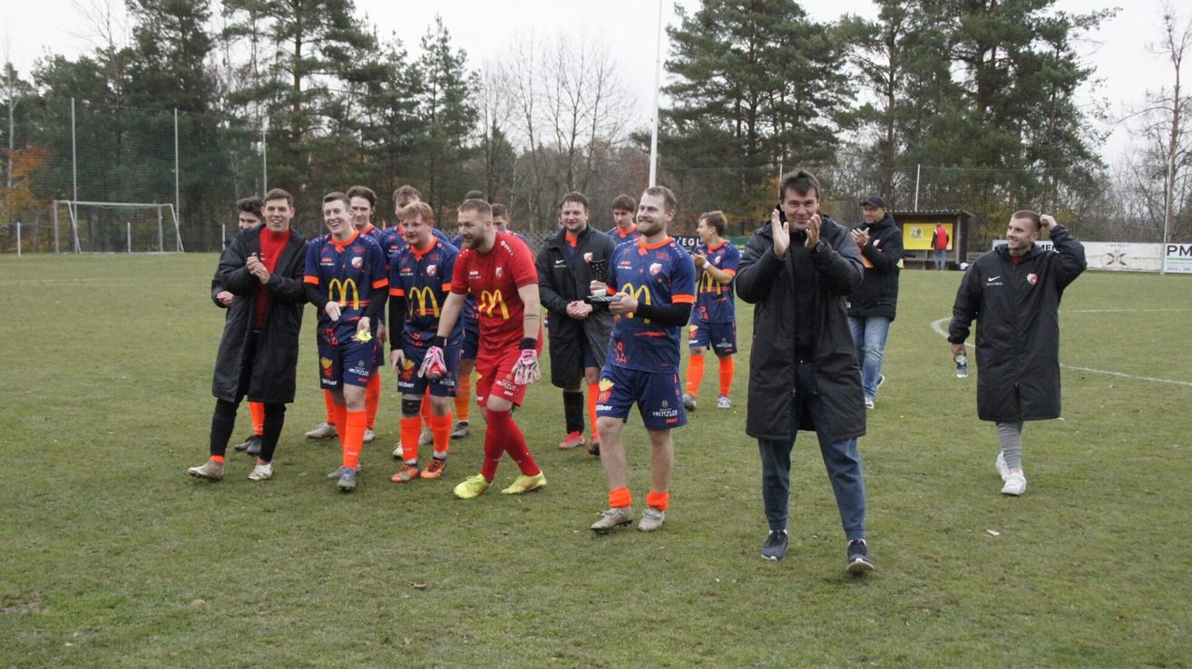 Eine Fußballmannschaft feiert auf einem Rasenfeld und trägt blaue und orangefarbene Trikots. Ein Spieler hält einen Pokal.