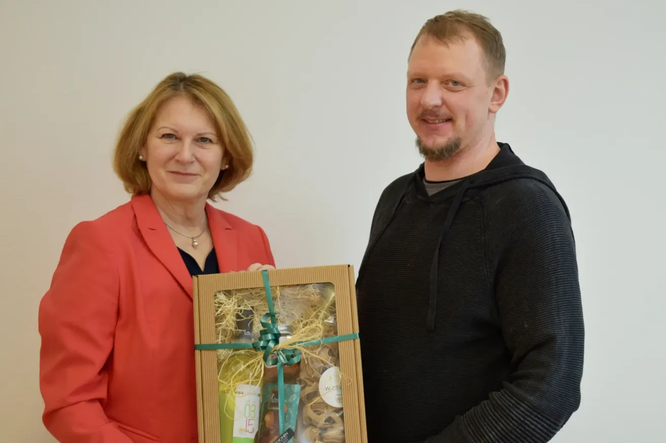 Eine Frau in einem orangefarbenen Blazer und ein Mann in einem schwarzen Hoodie stehen lächelnd zusammen und halten eine Geschenkbox mit grünem Band.