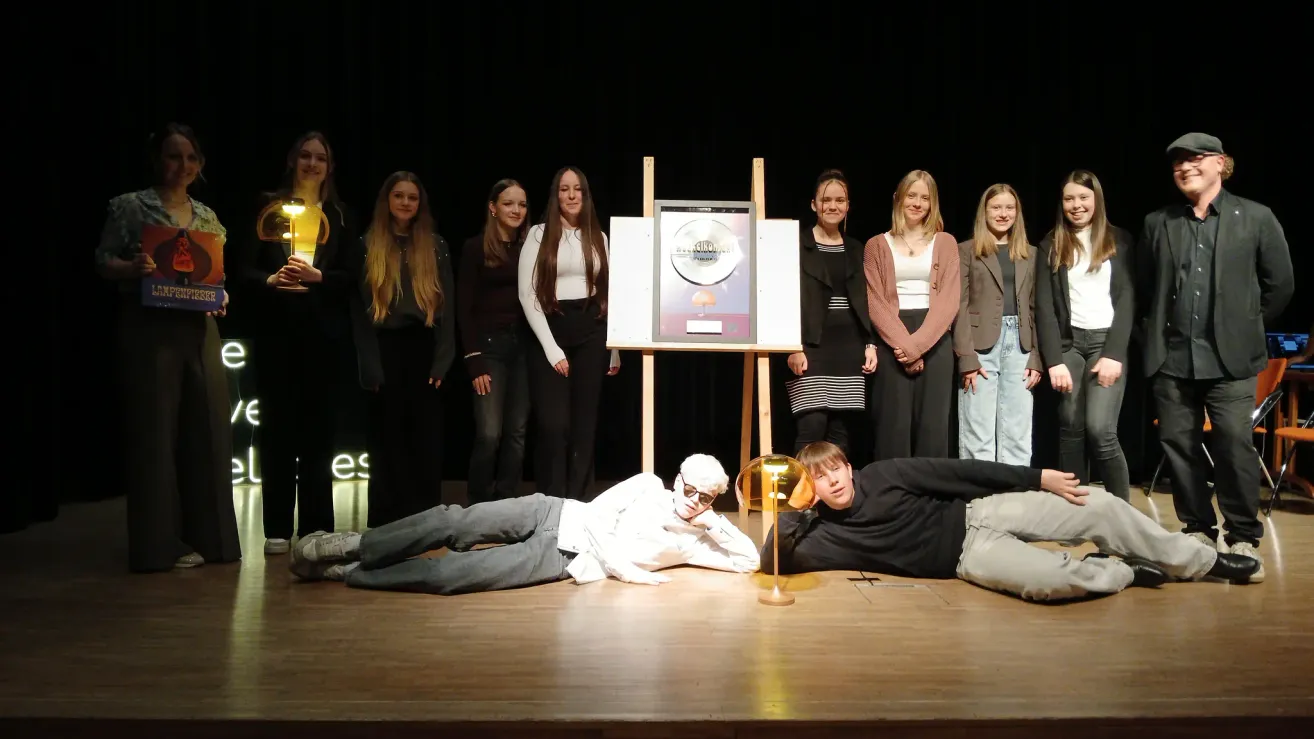 Eine Gruppe von Menschen steht auf der Bühne mit einem goldenen Preis in der Mitte. Einige halten Lichter, während zwei Menschen auf dem Boden liegen. Der Hintergrund ist dunkel.