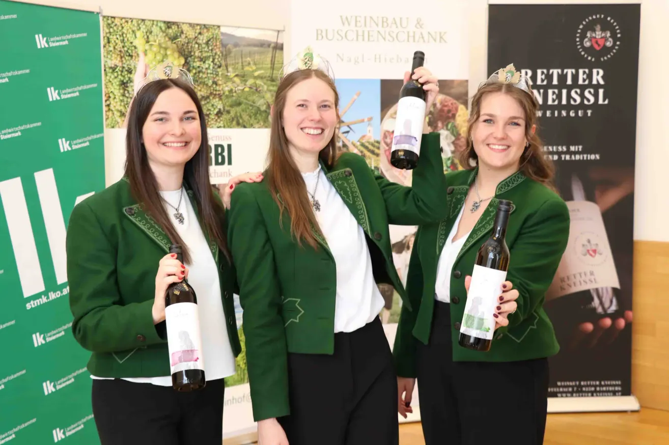 Drei Frauen in grünen Uniformen und weißen Hemden posieren für ein Foto, während sie Weinflaschen halten. Sie lächeln und stehen eng beieinander. Hinter ihnen ist ein Poster mit grünem Hintergrund und Text.