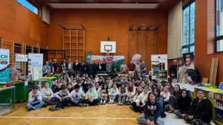 In einer Turnhalle versammeln sich eine große Gruppe von Schülern, Lehrern und Erwachsenen für eine Veranstaltung. Einige sitzen auf dem Boden, während andere hinter ihnen stehen. Ein Basketballkorb ist an der Wand montiert.