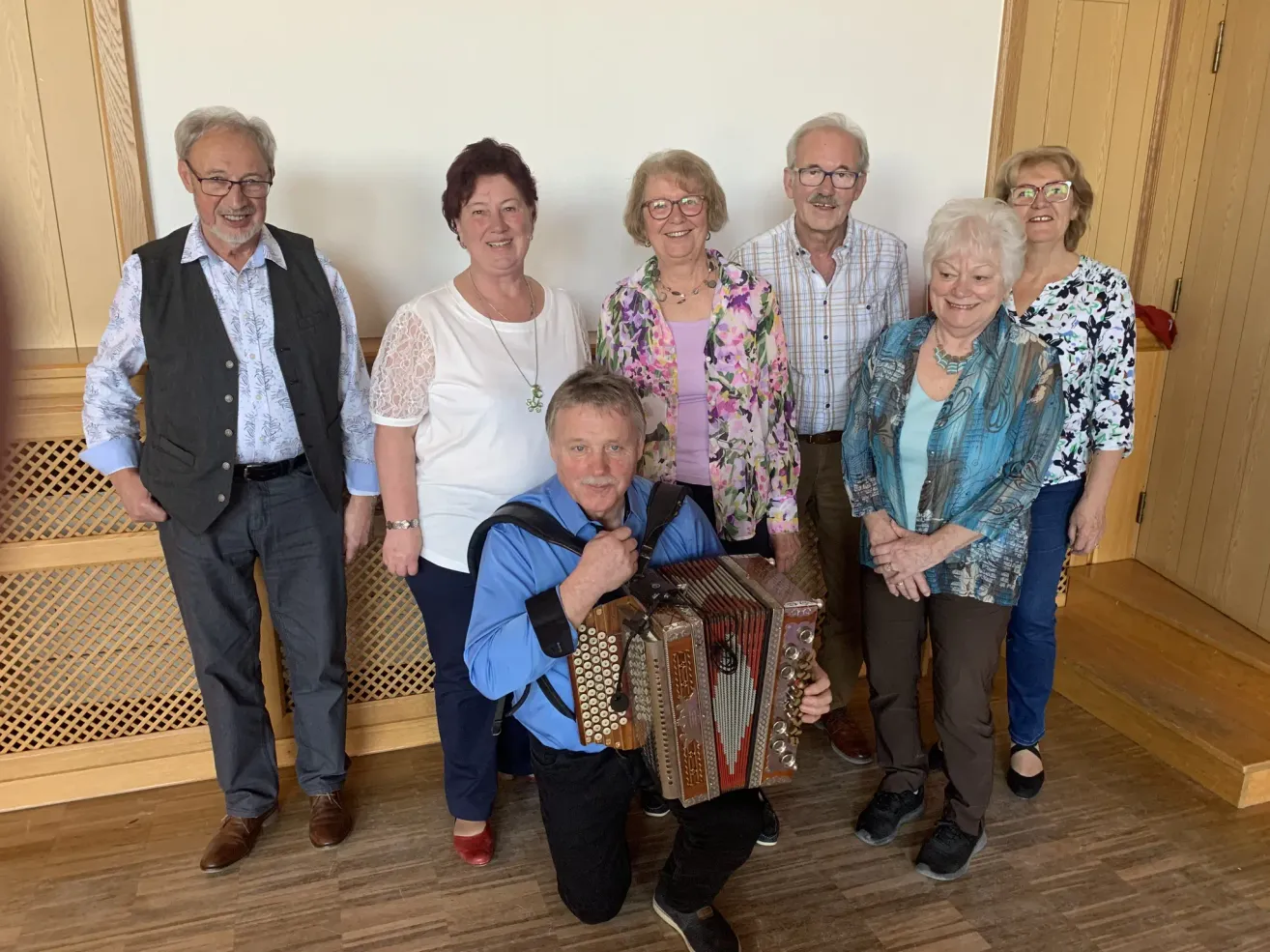 Eine Gruppe lächelnder älterer Erwachsener, darunter Männer und Frauen, steht in einem Raum zusammen. Ein Mann kniet und hält ein Akkordeon. Sie tragen alle Freizeitkleidung und scheinen für ein Foto zu posieren.