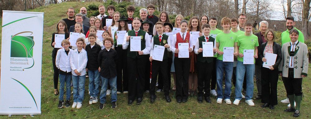 Eine Gruppe von Menschen steht auf einem Rasenfeld und hält Urkunden in der Hand. Sie alle lächeln und scheinen für ein Foto zu posieren.