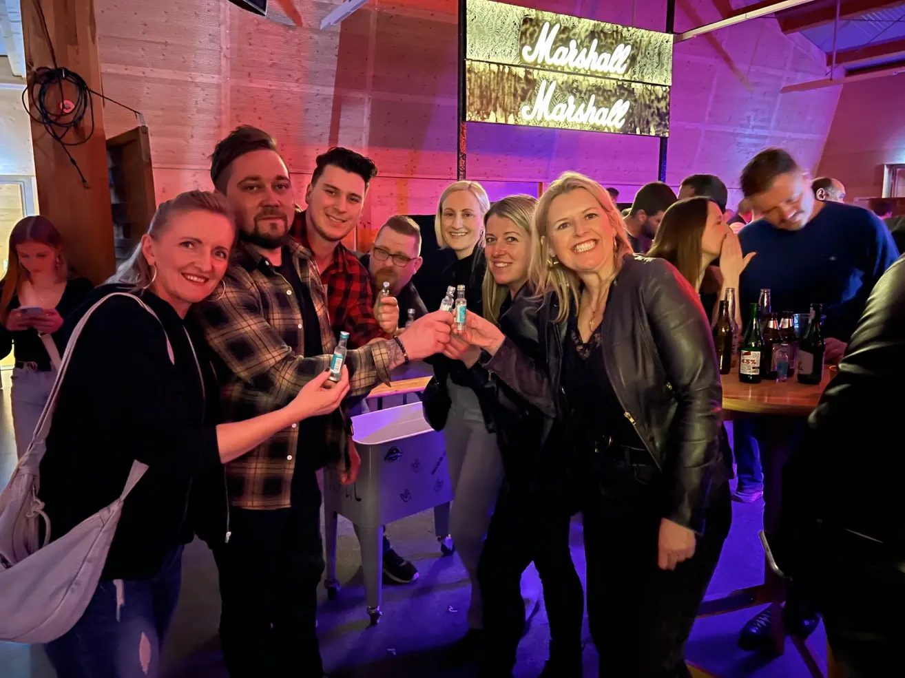 Eine Gruppe von Menschen versammelt sich drinnen und clinks ihre Gläser zusammen. Eine Frau in einer Lederjacke lächelt hell. Hinter ihnen steht ein Schild mit dem Wort Marshall. Im Hintergrund befinden sich Flaschen und ein Holztisch.