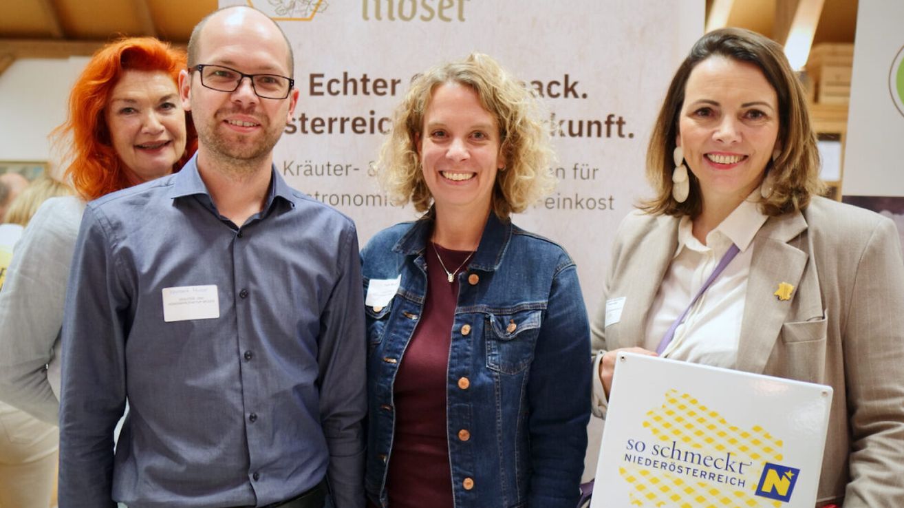 Drei Personen stehen vor einem Banner. Der Mann links trägt eine Brille und ein blaues Hemd, die Frau in der Mitte eine Jeansjacke und eine Halskette, und die Frau rechts hält ein Schild.