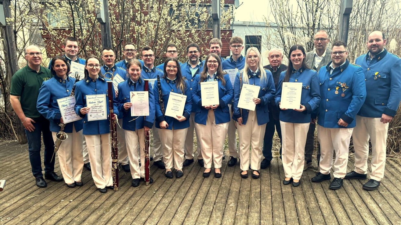 Eine Gruppe von Musikern in blauen Uniformen, die Urkunden halten, posiert für ein Foto auf einer Holzterrasse. Dahinter schmücken Bäume mit weißen Blüten die Gegend.