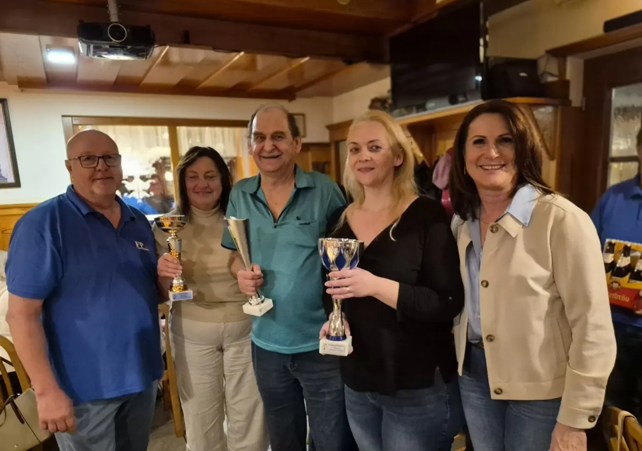 Fünf Menschen stehen lächelnd zusammen und halten Trophäen. Sie sind in einem Raum mit Holzdecke und Fernseher.