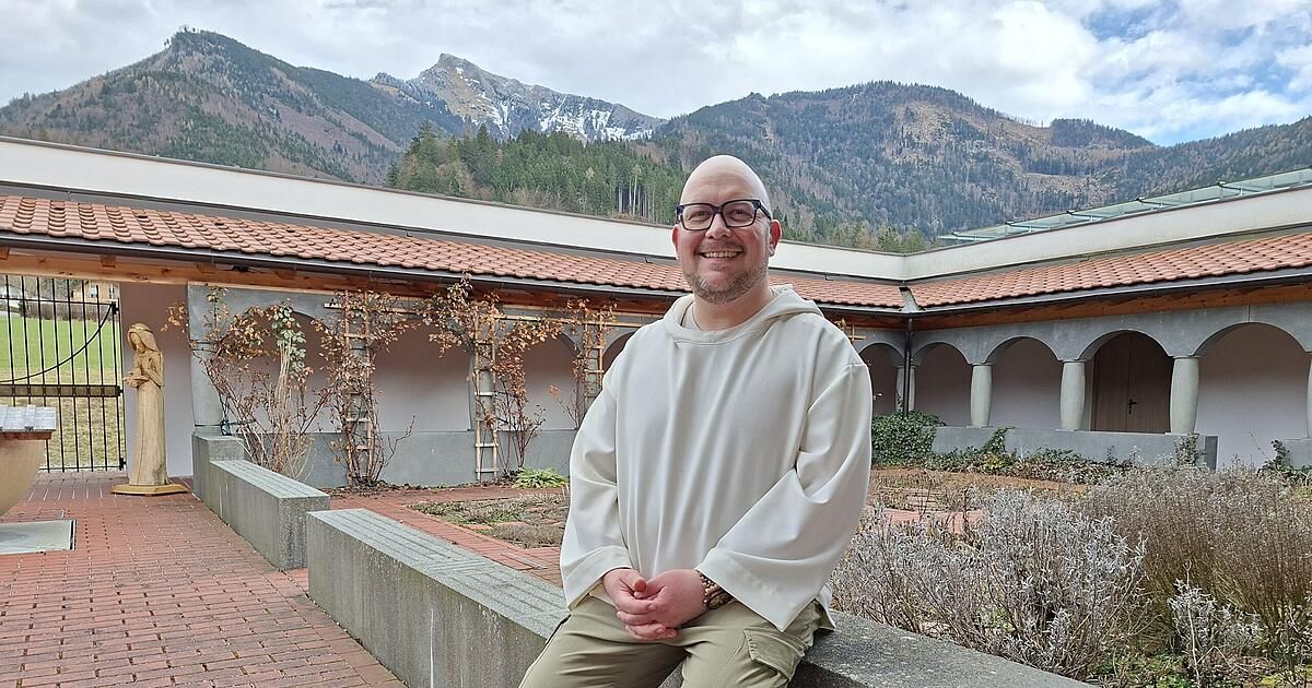 Ein lächelnder Mann in einer weißen Robe sitzt auf einer Steinbank vor einem Kloster mit Bergen im Hintergrund.