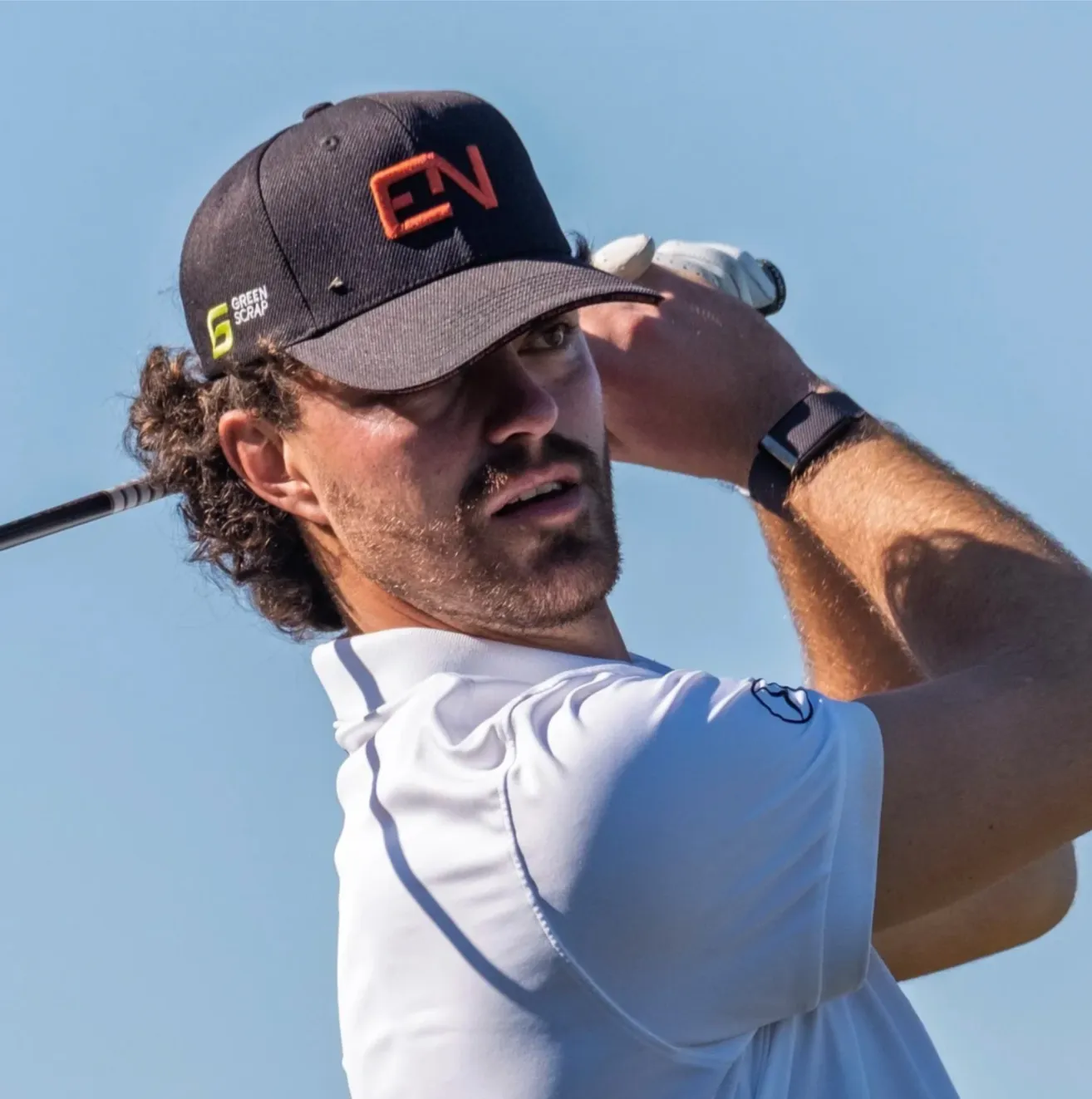 Ein Mann mit lockigem Haar, der eine Kappe und ein weißes Hemd trägt, schwingt einen Golfschläger. Er hält den Schläger in beiden Händen.