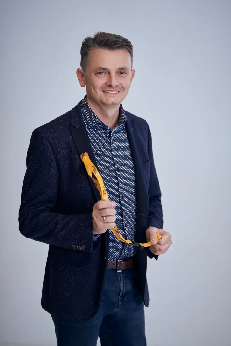 Ein Mann in einem dunkelblauen Mantel und weißem Hemd mit Punkten hält ein gelbes Namensschild in der Hand. Er steht vor einem weißen Hintergrund, lächelt und schaut in die Kamera.