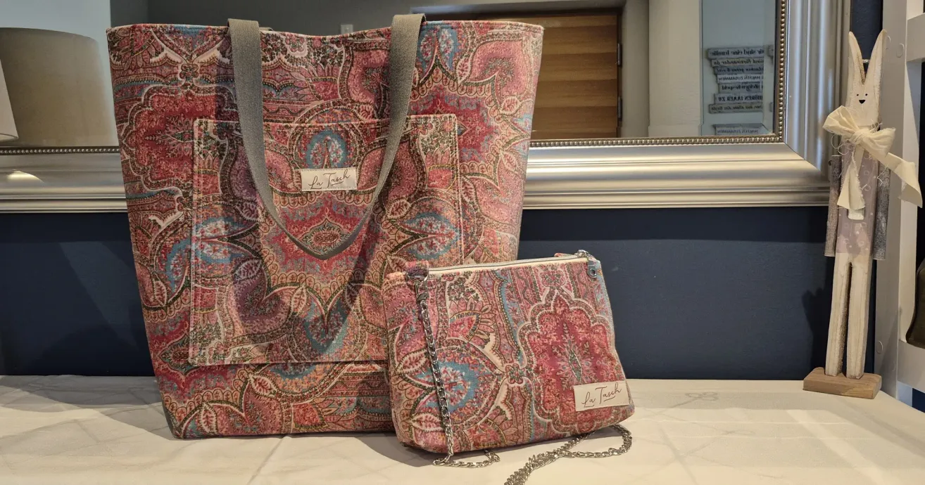 Zwei Taschen mit Paisleymuster und einem Etikett 'La Tuch' sind ausgestellt. Die größere Tasche ist eine Tragetasche mit Kettengriff, und die kleinere ist eine Handtasche mit Kettenriemen.