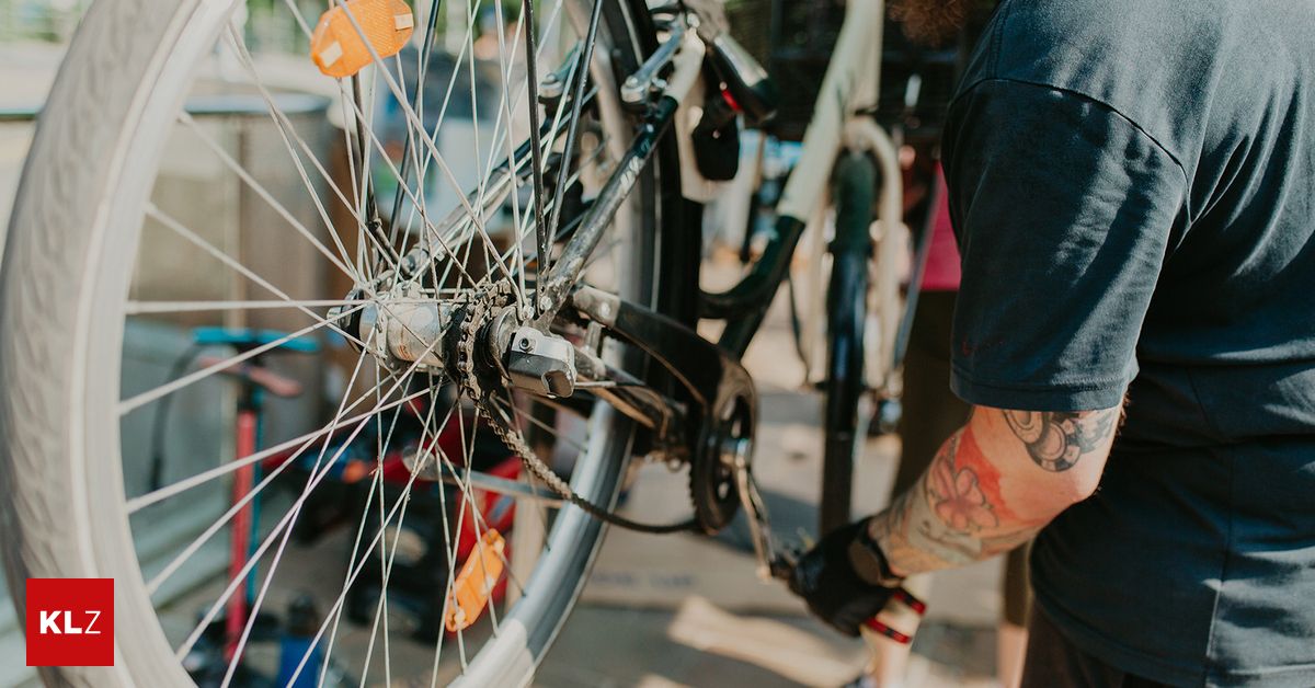 Eine Person in einem blauen Shirt arbeitet an einem Fahrrad und konzentriert sich auf das Hinterrad. Das Fahrrad hat mehrere orange Reflektoren, und die Person hat Tätowierungen an den Armen.