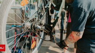 Eine Person in einem blauen Shirt arbeitet an einem Fahrrad und konzentriert sich auf das Hinterrad. Das Fahrrad hat mehrere orange Reflektoren, und die Person hat Tätowierungen an den Armen.