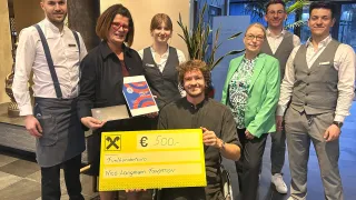 Ein Mann im Rollstuhl hält einen großen gelben Scheck, während eine Gruppe von Menschen hinter ihm steht. Sie tragen alle formelle Kleidung.