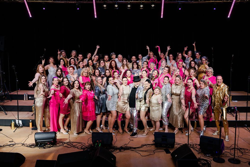 Eine große Gruppe von Menschen in pinken und silbernen Kostümen posiert für ein Foto auf einer Bühne mit Lautsprechern und Lichtern im Hintergrund.