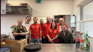 Eine Gruppe von Menschen in roten Hemden posiert für ein Foto in einer Küche. Einer hält eine Schüssel, während ein anderer eine Halskette trägt.