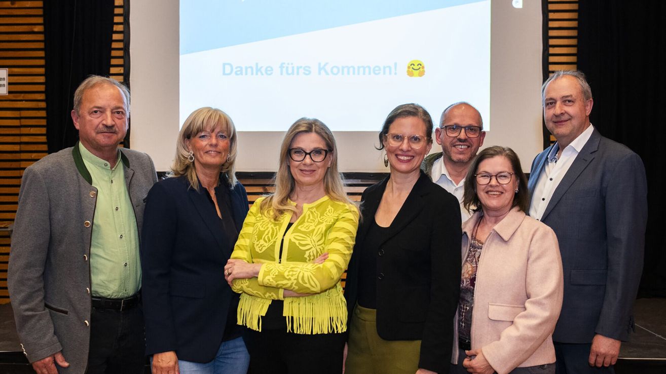 Fünf lächelnde Personen posieren vor einer Projektionsleinwand mit der Botschaft 'Danke fürs Kommen!'. Die Frau in Gelb trägt ein Top mit Fransen, die anderen tragen Brillen.
