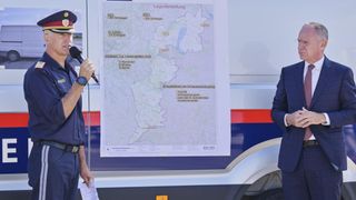 Ein uniformierter Beamter spricht in ein Mikrofon und hält eine große Karte mit Orten und Statistiken. Ein weiterer Mann im Anzug steht hinter der Karte.