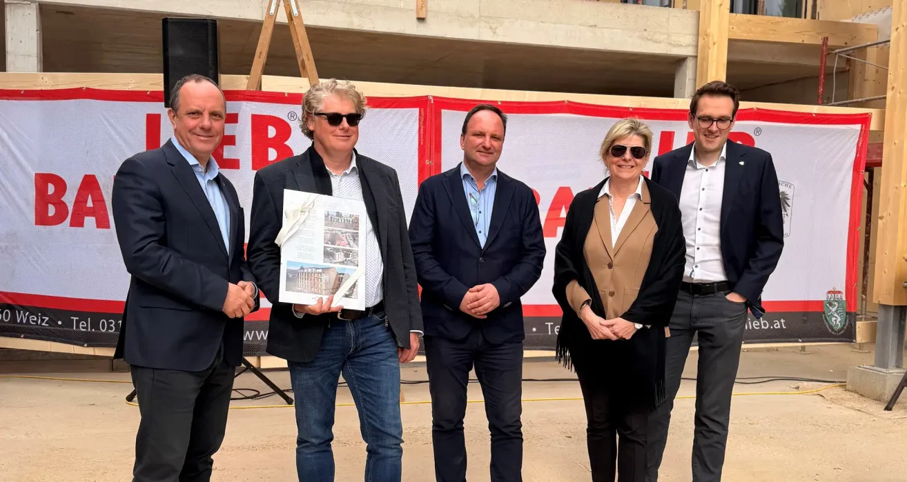 Fünf Menschen, alle formell gekleidet, stehen vor einer Baustelle. Ein Mann hält ein Buch. Ein anderer Mann hat eine Sonnenbrille. Eine Frau hält ein Stück Papier.