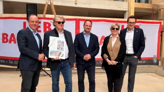 Fünf Menschen, alle formell gekleidet, stehen vor einer Baustelle. Ein Mann hält ein Buch. Ein anderer Mann hat eine Sonnenbrille. Eine Frau hält ein Stück Papier.