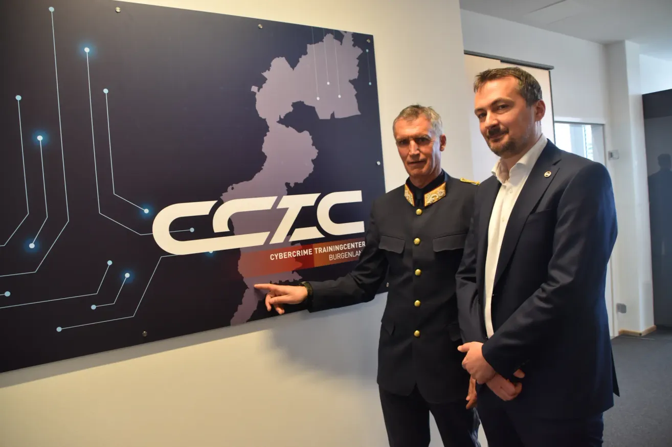 Zwei Männer stehen vor einem Schild des Cybercrime Training Centers in Burgenland, Deutschland. Ein Mann in Uniform zeigt auf das Schild, während der andere zusieht.