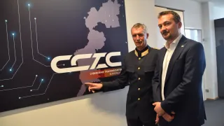 Zwei Männer stehen vor einem Schild des Cybercrime Training Centers in Burgenland, Deutschland. Ein Mann in Uniform zeigt auf das Schild, während der andere zusieht.