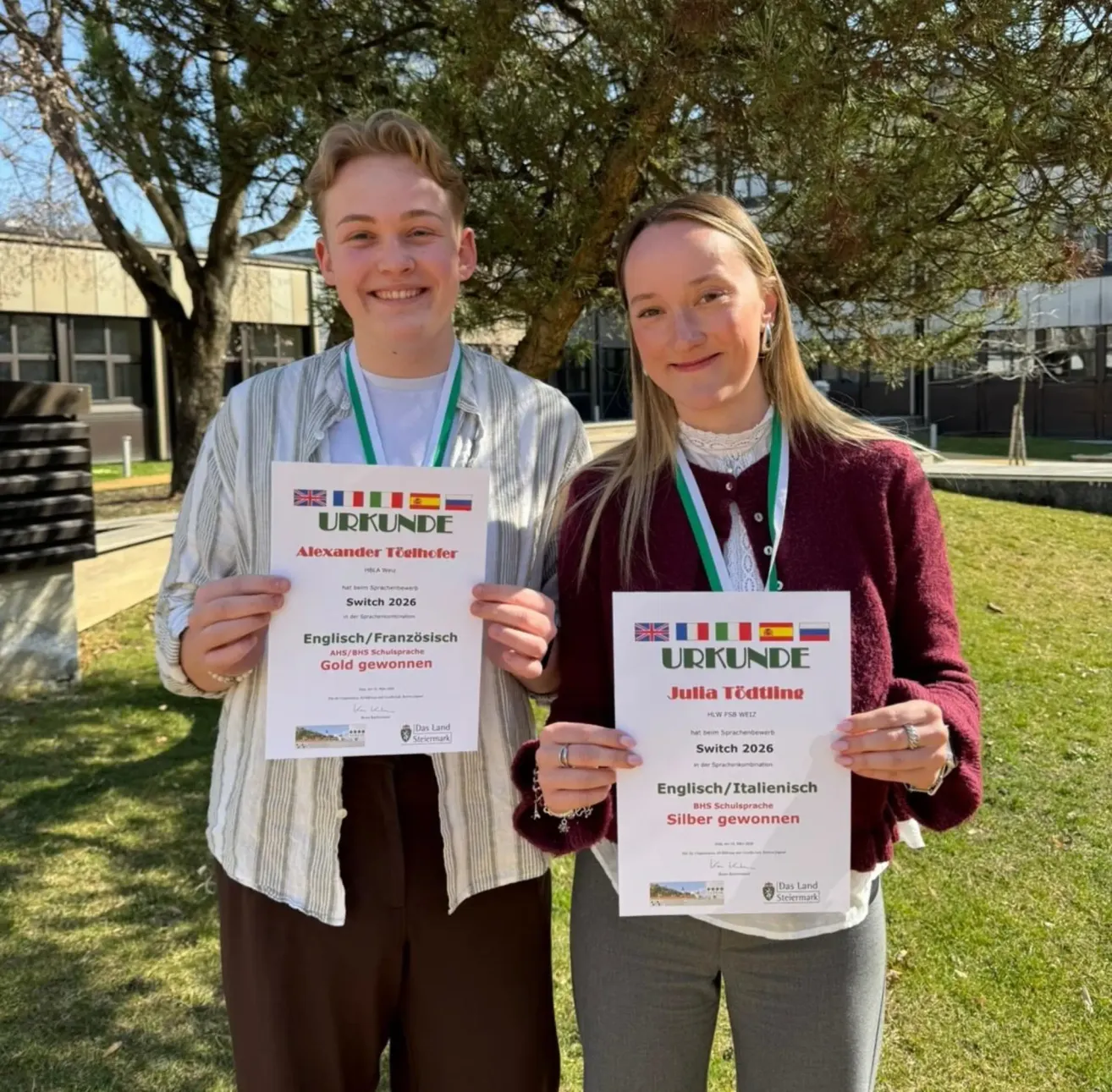 Zwei Personen posieren mit Urkunden im Freien. Der Junge hält ein goldenes Siegerzertifikat, die Mädchen ein silbernes. Beide tragen Medaillen und stehen unter einem Baum. Dahinter sind ein Gebäude und ein Rasenbereich zu sehen.