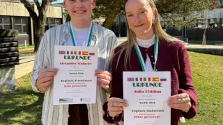 Zwei Personen posieren mit Urkunden im Freien. Der Junge hält ein goldenes Siegerzertifikat, die Mädchen ein silbernes. Beide tragen Medaillen und stehen unter einem Baum. Dahinter sind ein Gebäude und ein Rasenbereich zu sehen.