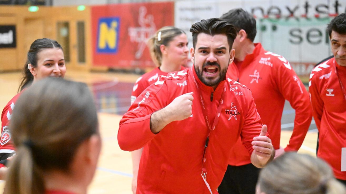 Ein Mann in einem roten Jackett zeigt mit seinem erhobenen Daumen und geballter Faust auf einem Sportplatz. Andere Spieler in roten Trikots sind im Hintergrund.