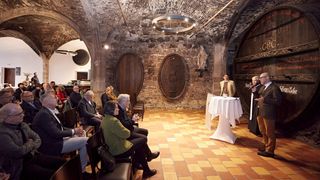 Eine Gruppe von Menschen sitzt in einem Raum mit Steinwänden und gefliesten Böden. Ein Mann steht an einem Tisch. Zwei große Holzfässer und eine Statue befinden sich an der Wand.