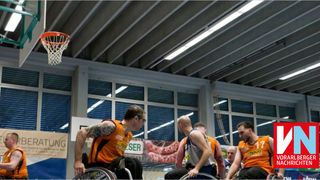 Vier Männer im Rollstuhl spielen Basketball in einer Halle. Zwei von ihnen schauen nach links.