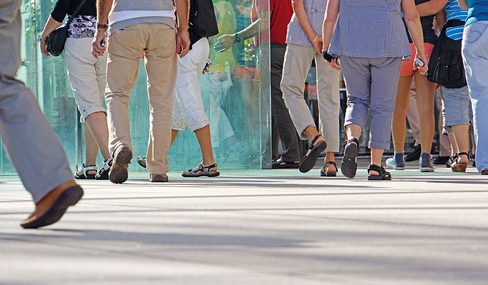 Mehrere Menschen gehen in einer Reihe auf einem Bürgersteig, hinter ihnen steht eine Glasbarriere. Sie tragen Freizeitkleidung und Sandalen.