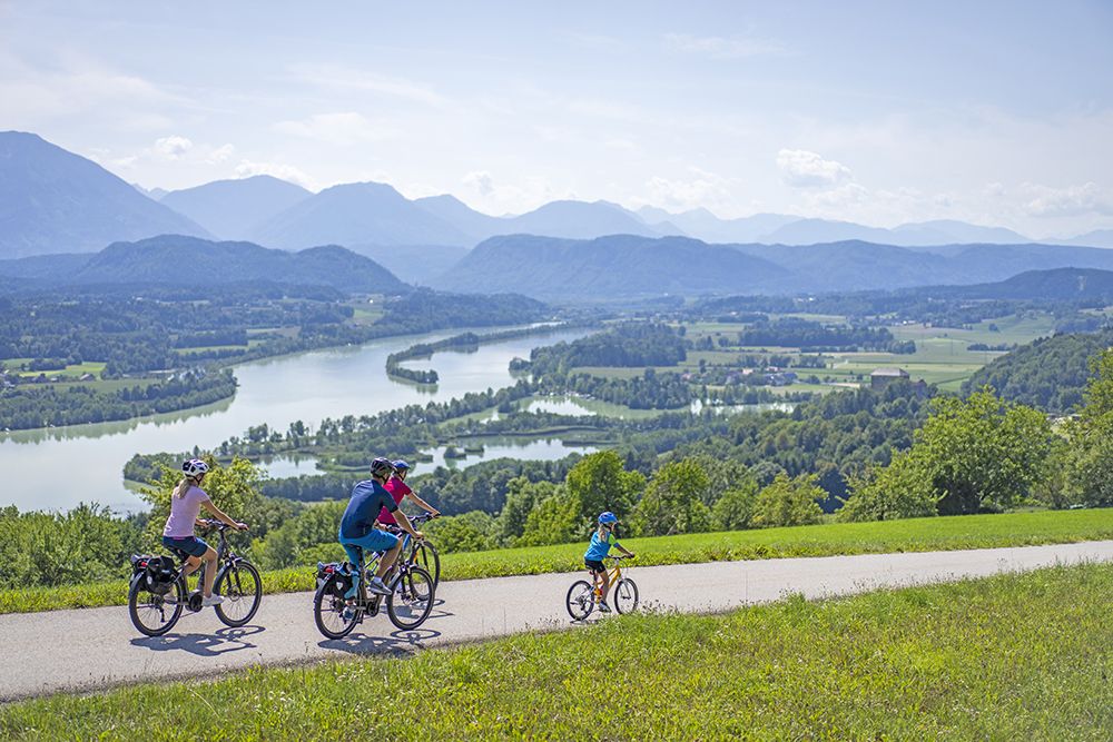 Vier Personen fahren auf einem Pfad mit Fahrrädern und genießen einen schönen Tag. Sie haben einen Blick auf einen Fluss und Berge in der Ferne.