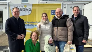 Eine Familie und mehrere Erwachsene posieren für ein Foto vor einem Gebäude. Ein Mädchen hält ein Kuscheltier und lächelt.