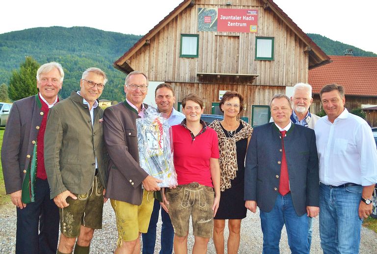 Eine Gruppe von Menschen steht vor einem Holzgebäude mit einem Schild, das Jauntaler Hadn Zentrum liest. Sie sind alle in traditioneller Kleidung gekleidet und lächeln. Die Frau in der Mitte hält einen Blumenstrauß.