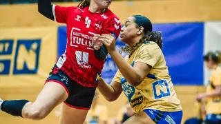 Zwei Frauen spielen Handball. Eine springt, um einen roten Ball zu werfen, während die andere sie blockt. Das Publikum beobachtet im Hintergrund.
