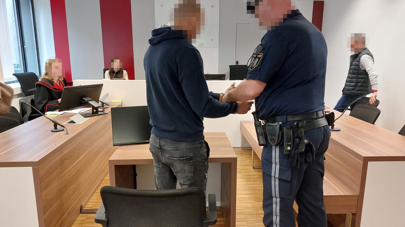 Eine Person in einer blauen Kapuzenjacke wird von einem Polizisten in einem Gerichtssaal eskortiert. Sie schütteln Hände.