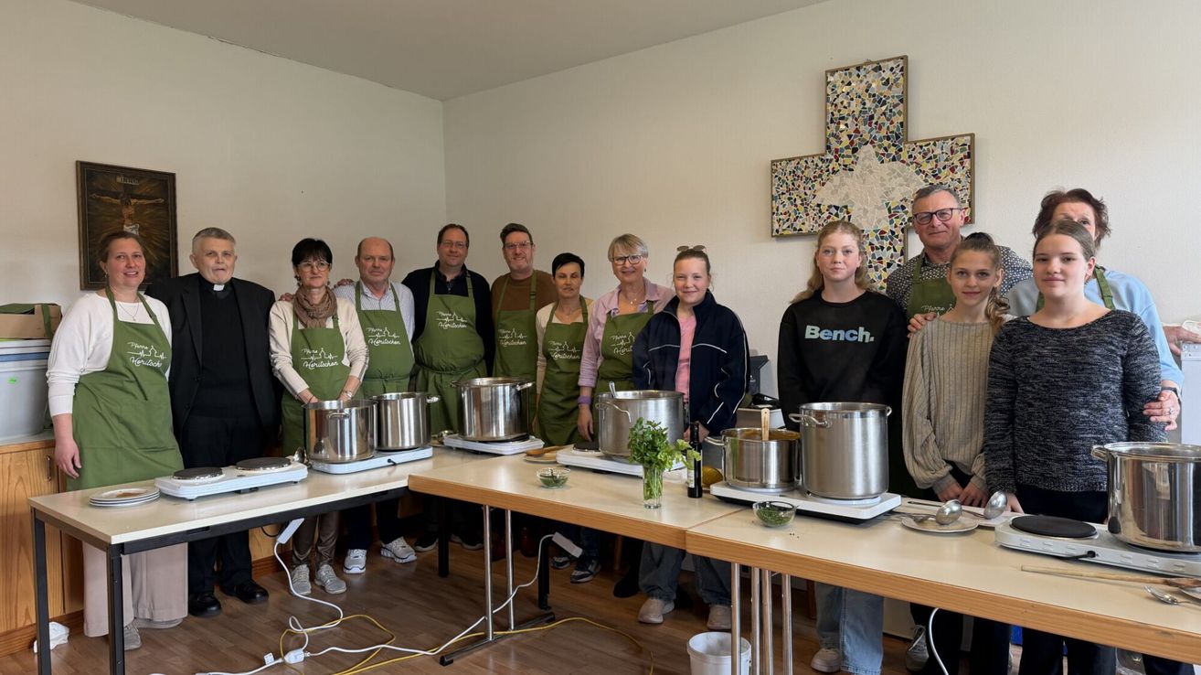 Eine Gruppe von Menschen in grünen Schürzen steht um einen Küchentisch mit Töpfen und Flaschen. Eine Frau in einem schwarzen Shirt hält eine Flasche.