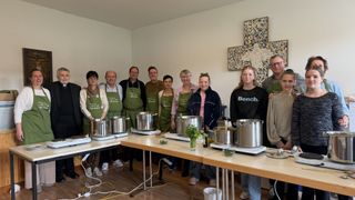 Eine Gruppe von Menschen in grünen Schürzen steht um einen Küchentisch mit Töpfen und Flaschen. Eine Frau in einem schwarzen Shirt hält eine Flasche.