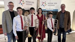 Eine Gruppe junger Musiker, bestehend aus Jungen und einem Mädchen, posiert mit Posaunen vor einem Banner mit der Aufschrift 'Musik der Jugend'.