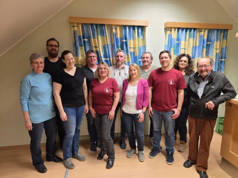 Eine Gruppe von Menschen in Freizeitkleidung steht zusammen in einem Raum, wahrscheinlich für ein Gruppenfoto. Sie lächeln alle und scheinen glücklich zu sein. Der Raum hat Holzböden und weiße Wände mit blauen Vorhängen.