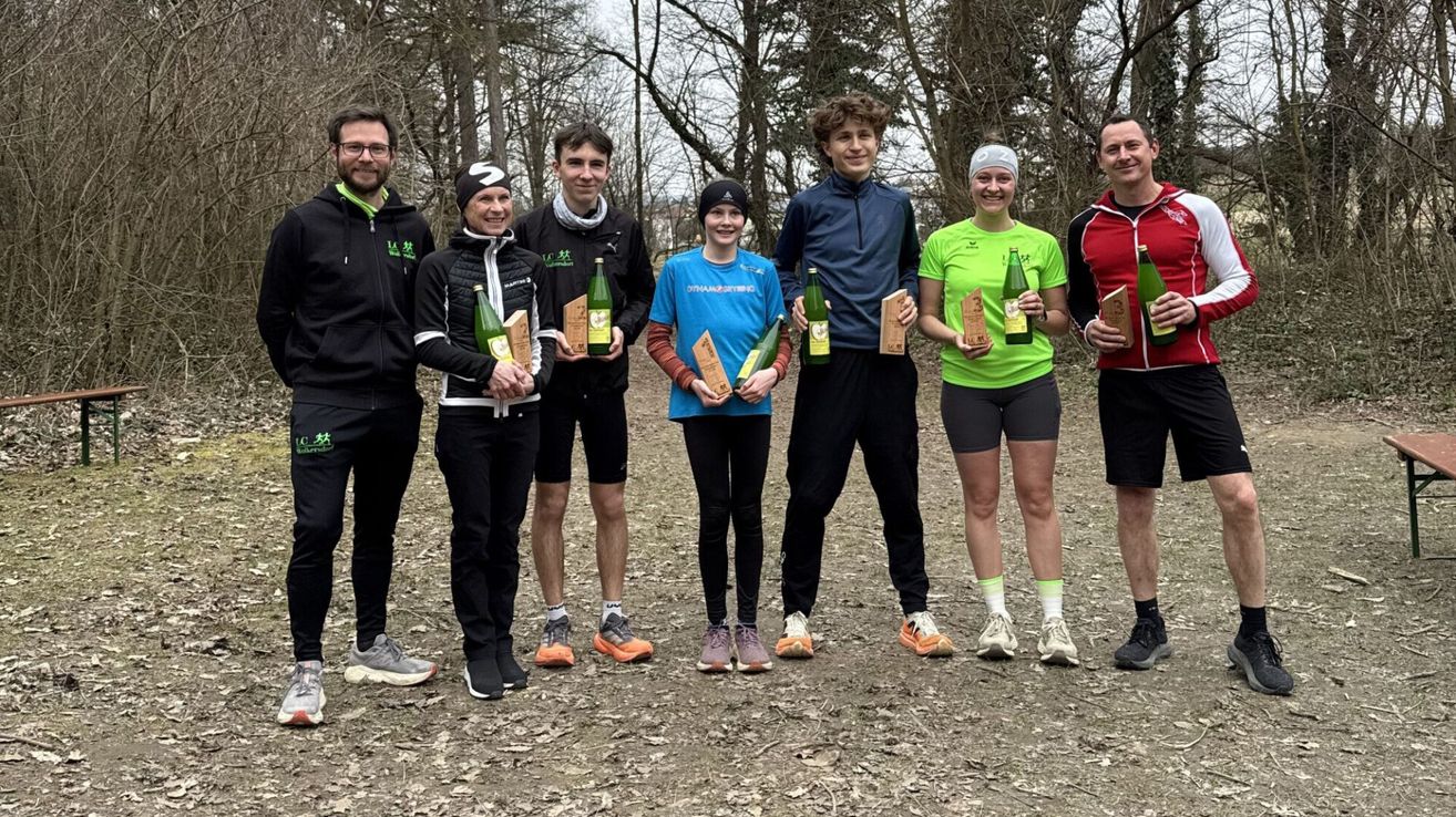 Eine Gruppe von Menschen im Wald posiert für ein Foto. Sie halten Flaschen und Auszeichnungen. Sie sind in verschiedene Kleidungsstücke gekleidet, darunter Jacken, Sneaker und Mützen.