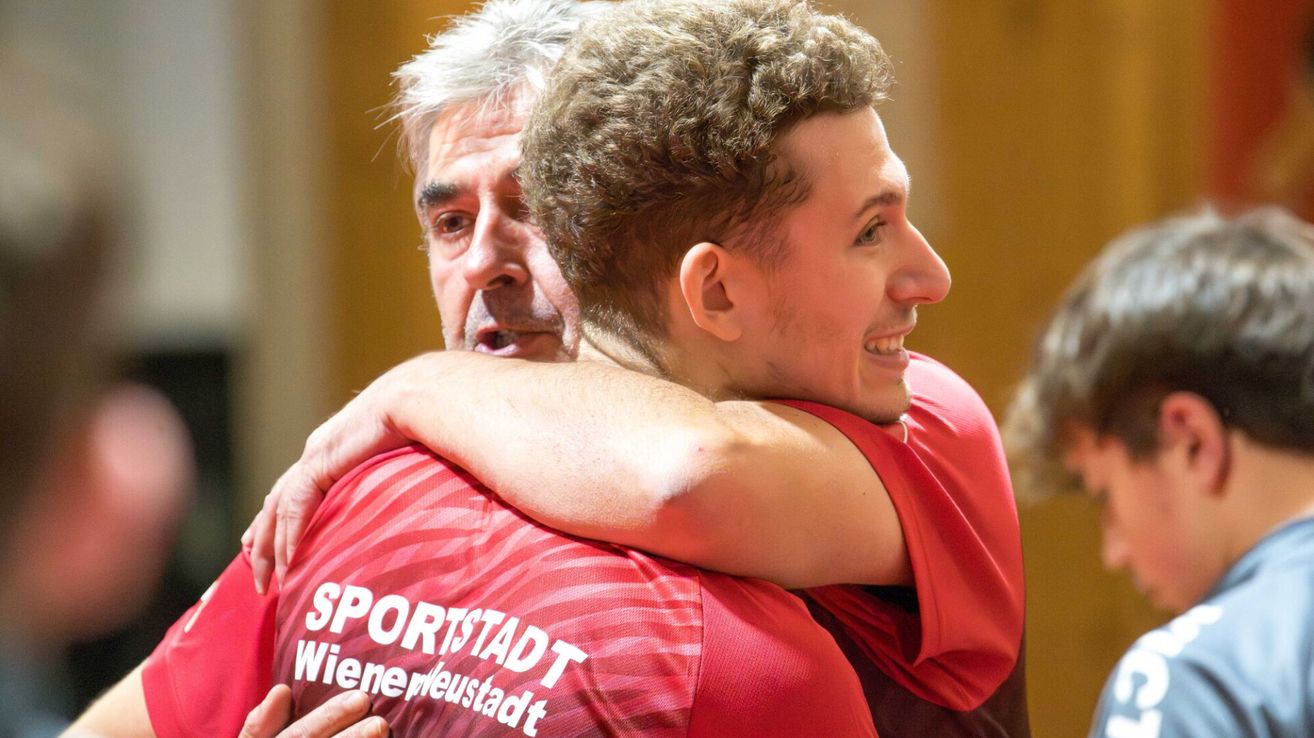 Zwei Männer umarmen sich. Einer trägt ein rotes Sporthemd mit dem Text 'SPORTSTADT Wieneuestadt.' Sie scheinen zu feiern.