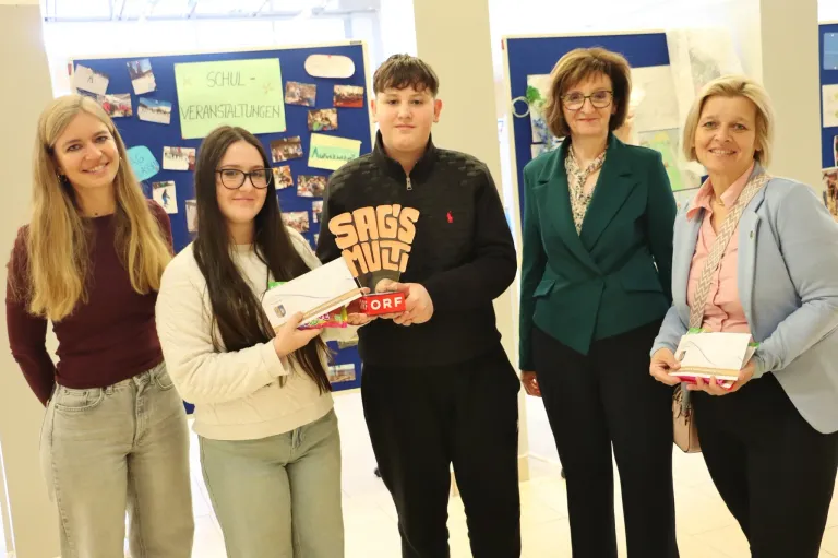 Ein junges Mädchen mit Brille hält eine Schachtel mit Süßigkeiten und steht neben einem jungen Mann mit einer Jacke, auf der Sag's Muli steht, während eine Frau in einem grünen Blazer lächelt. Sie sind in einer Schule mit Bulletins an den Wänden.