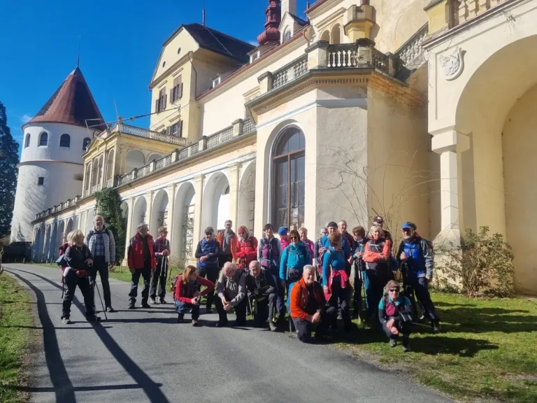 Eine Gruppe von Wanderern in Jacken und Mützen posiert für ein Foto vor einem alten Gebäude. Einige knien, andere stehen und halten Wanderstöcke.
