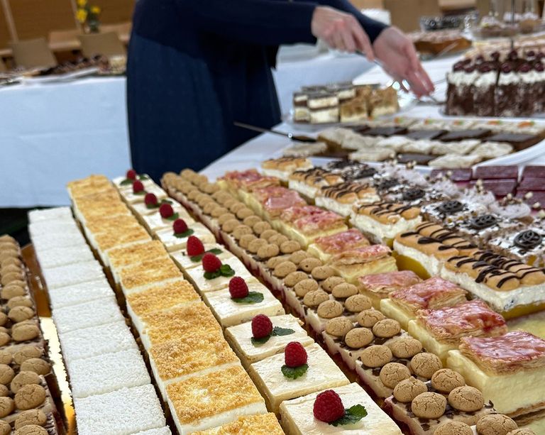 Ein Tisch ist mit einer Vielzahl von Desserts gefüllt, darunter Kuchen, Törtchen und Käsekuchen, garniert mit Himbeeren und anderen Toppings. Eine Person in einem blauen Kleid arrangiert die Desserts.