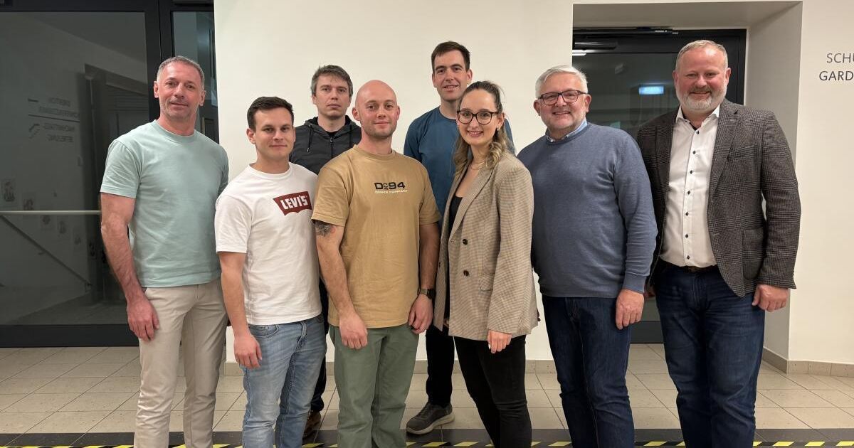 Eine Gruppe von Menschen, wahrscheinlich Kollegen, steht in einem modernen Raum, lächelt und schaut zur Kamera. Eine Frau in der Mitte trägt eine Brille und einen Blazer. Der Hintergrund zeigt eine weiße Wand und eine Glastür.