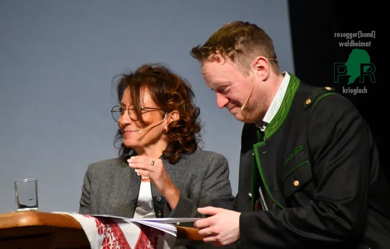 Ein Mann und eine Frau stehen an einem Podium, wahrscheinlich während einer Preisverleihung. Die Frau hält ein Papier in der Hand, während der Mann lächelt und sie ansieht. Beide haben Mikrofone an den Ohren.