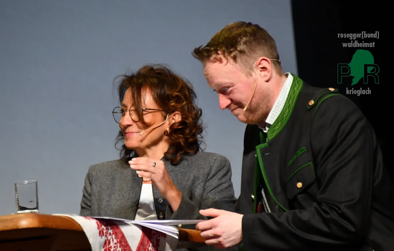 Ein Mann und eine Frau stehen an einem Podium, wahrscheinlich während einer Preisverleihung. Die Frau hält ein Papier in der Hand, während der Mann lächelt und sie ansieht. Beide haben Mikrofone an den Ohren.