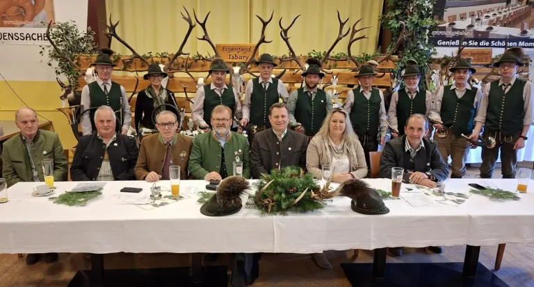 Eine Gruppe von Menschen in traditioneller Kleidung posiert für ein Foto an einem Tisch mit Biergläsern und Geweihen. Sie stehen vor einer Holzwand mit einem Schild, das 'Eigenjagd Isbary' steht.